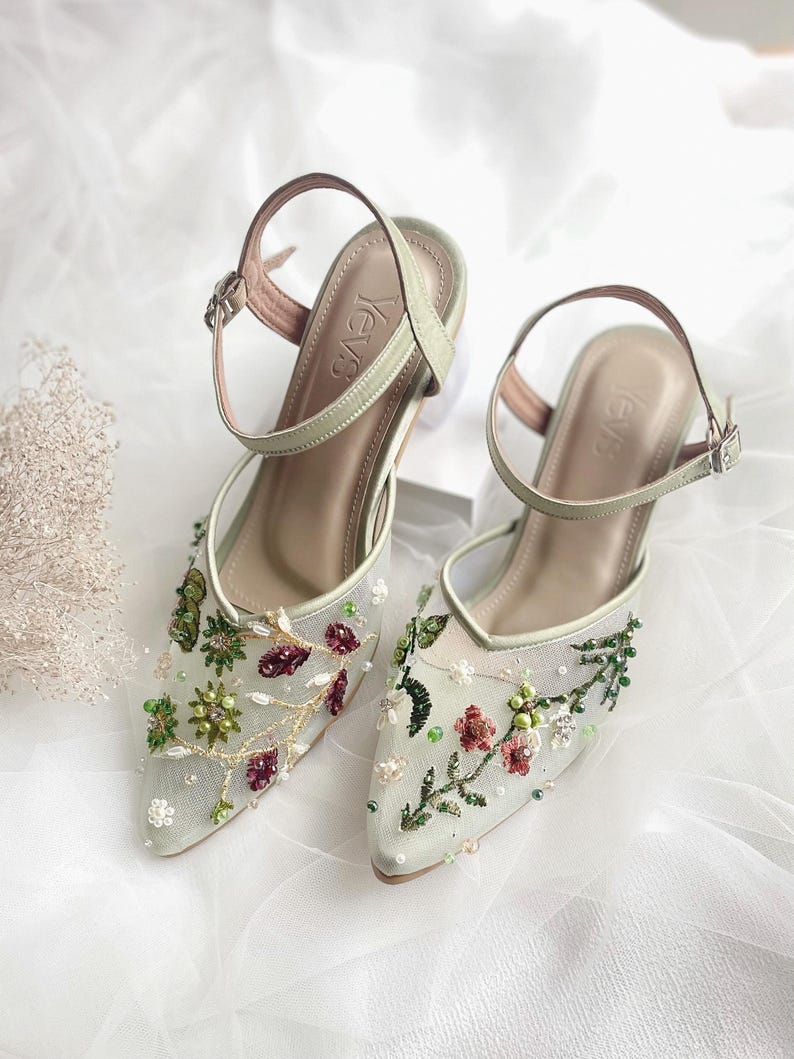 K&ouml;nnte beinhalten: Ein Paar hellgr&uuml;ne High-Heel-Schuhe mit zarter Blumenstickerei und einem Mesh-Gewebe. Die Schuhe haben eine spitze Zehenpartie und einen Kn&ouml;chelriemen mit einer Schnalle. Der Markenname "Yev's" ist auf der Innenseite der Schuhe sichtbar.