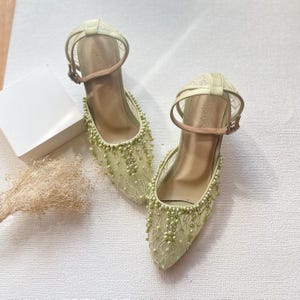 May include: A pair of light green, heeled shoes with ankle straps. The shoes feature a delicate lace overlay and are embellished with small, light green beads. The heels are approximately 2 inches high. The straps are a light brown color.