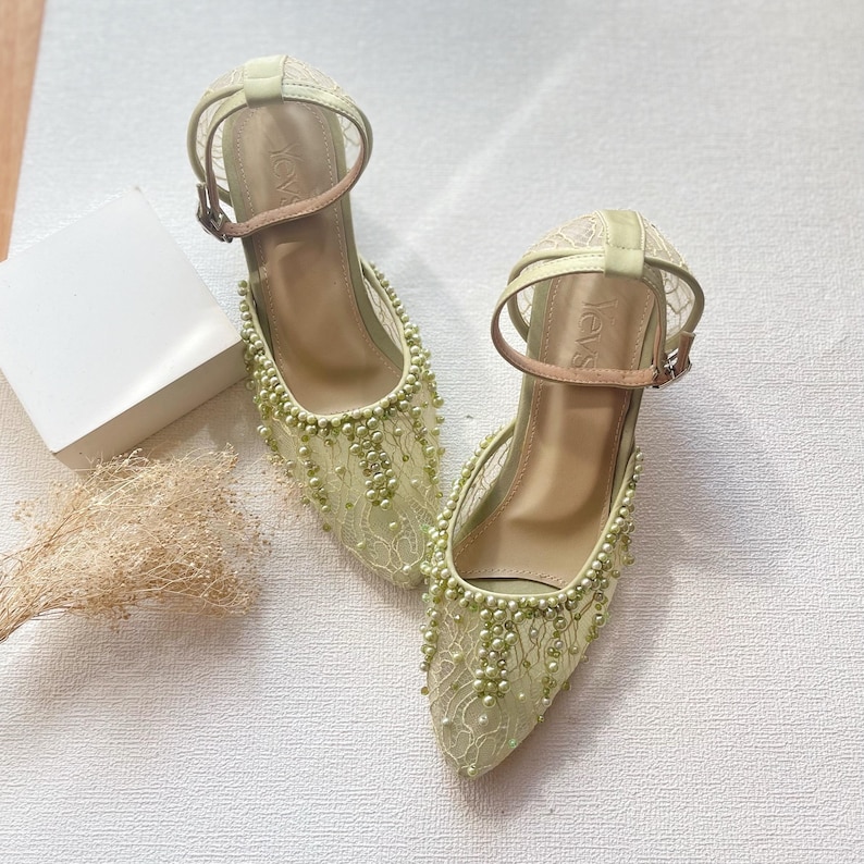 May include: A pair of light green, heeled shoes with ankle straps. The shoes feature a delicate lace overlay and are embellished with small, light green beads. The heels are approximately 2 inches high. The straps are a light brown color.