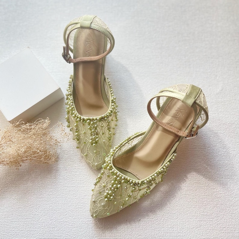 May include: A pair of light green, pointed-toe heels with ankle straps. The shoes are adorned with delicate lace and small, light green beads. The heels have a medium height and a buckle closure. The overall design is elegant and suitable for special occasions.