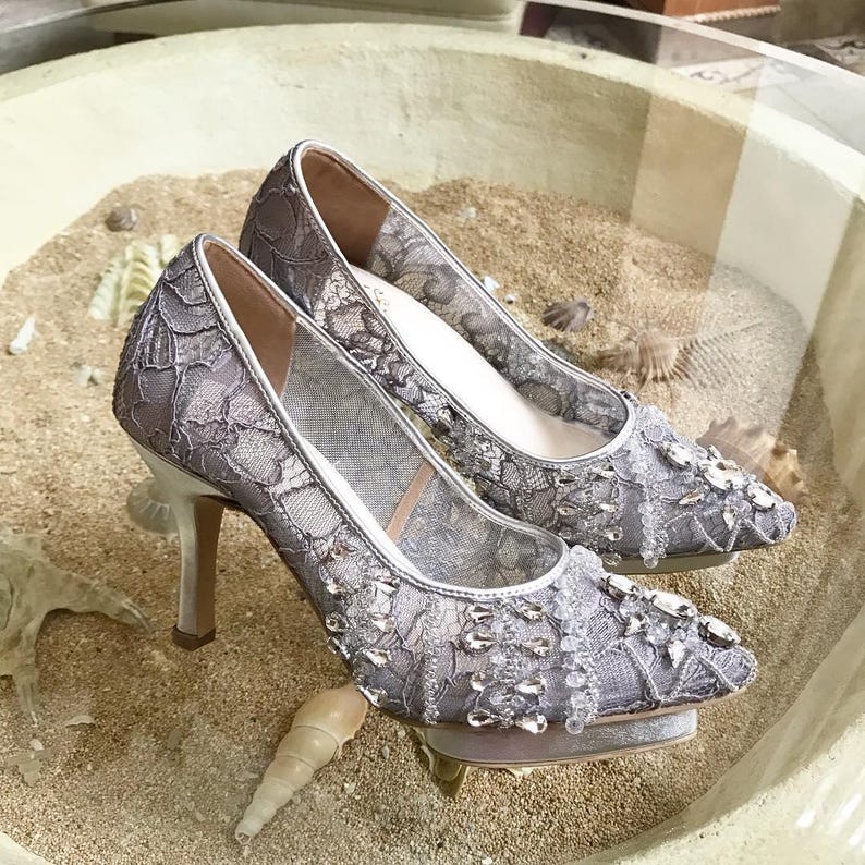 May include: A pair of silver high-heeled shoes with lace and beaded embellishments. The shoes are on a sandy surface.