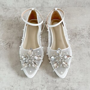 May include: A pair of white pointed-toe flats featuring a sheer mesh upper adorned with scattered small white pearls. Each shoe has a prominent floral-shaped cluster of clear faceted crystals at the toe and a thin white ankle strap with a buckle.