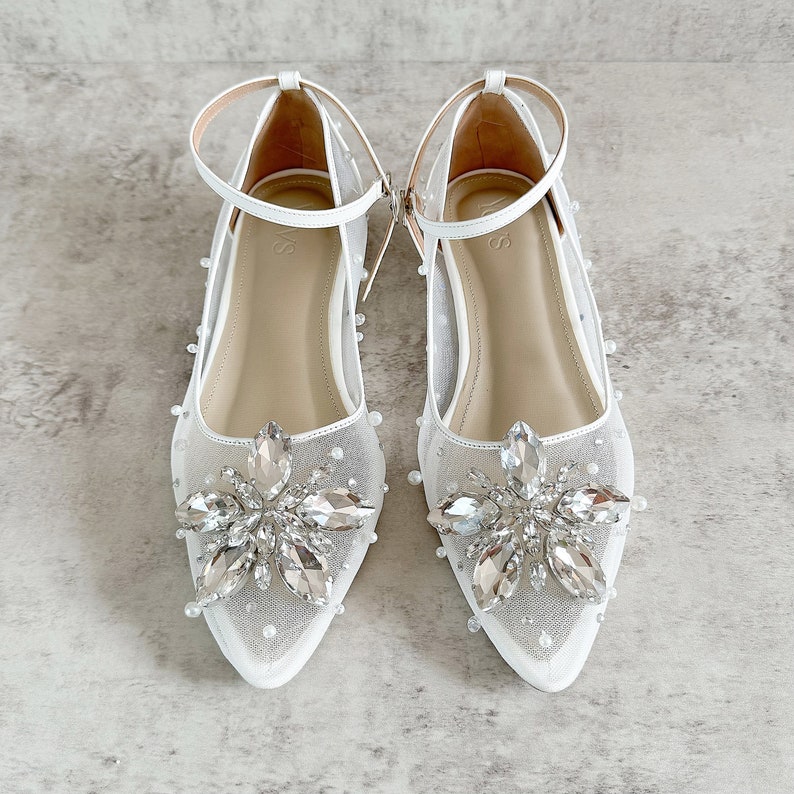 May include: A pair of white pointed-toe flats featuring a sheer mesh upper adorned with scattered small white pearls. Each shoe has a prominent floral-shaped cluster of clear faceted crystals at the toe and a thin white ankle strap with a buckle.