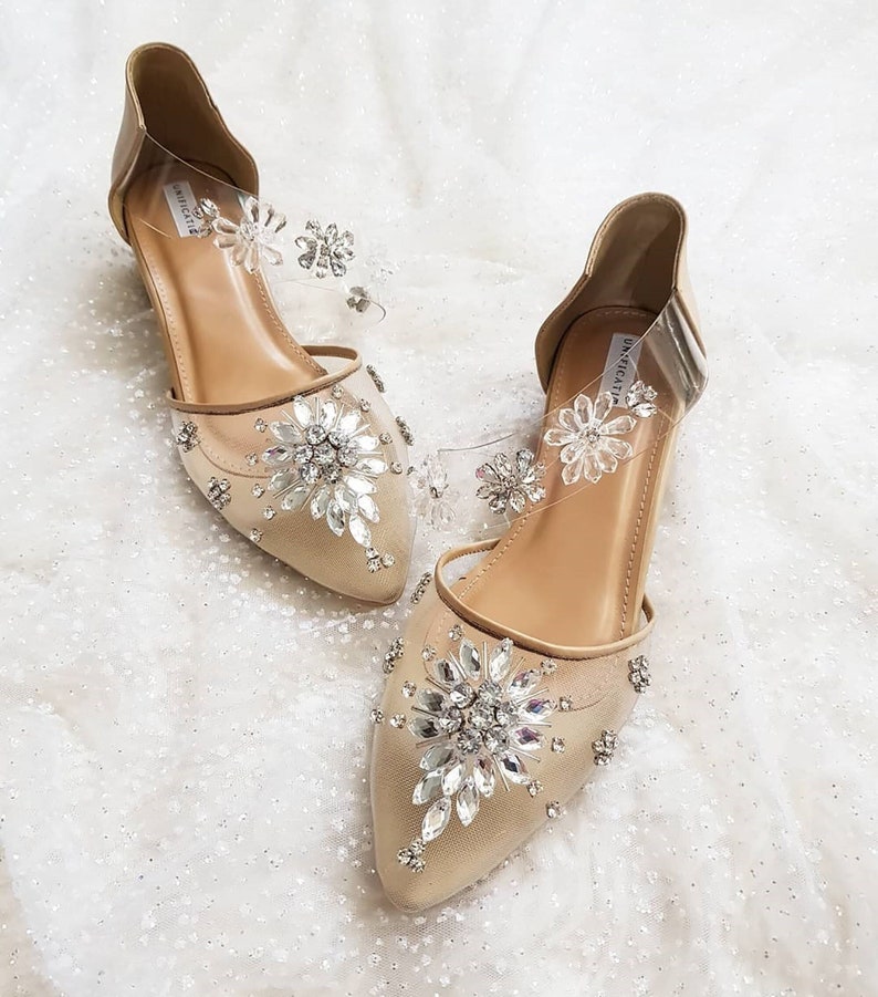 Puede incluir: Un par de zapatos de novia beige con punta puntiaguda y un delicado tejido de malla. Los zapatos est&aacute;n adornados con grandes diamantes de imitaci&oacute;n brillantes en un dise&ntilde;o floral. Los zapatos tienen una correa transparente con peque&ntilde;os diamantes de imitaci&oacute;n.