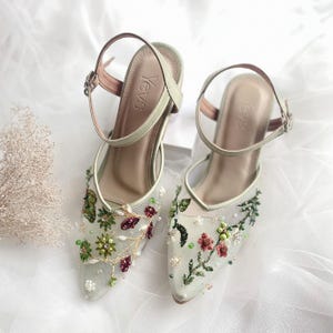 K&ouml;nnte beinhalten: Ein Paar hochhackige Schuhe in Salbeigr&uuml;n mit zarter Blumenstickerei und Perlenakzenten. Die Schuhe haben eine spitze Zehenpartie und einen Kn&ouml;chelriemen mit einer Schnalle. Der Markenname "YevS" ist auf der R&uuml;ckseite der Schuhe sichtbar.