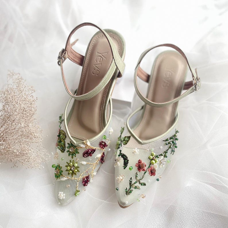 K&ouml;nnte beinhalten: Ein Paar hochhackige Schuhe in Salbeigr&uuml;n mit zarter Blumenstickerei und Perlenakzenten. Die Schuhe haben eine spitze Zehenpartie und einen Kn&ouml;chelriemen mit einer Schnalle. Der Markenname "YevS" ist auf der R&uuml;ckseite der Schuhe sichtbar.