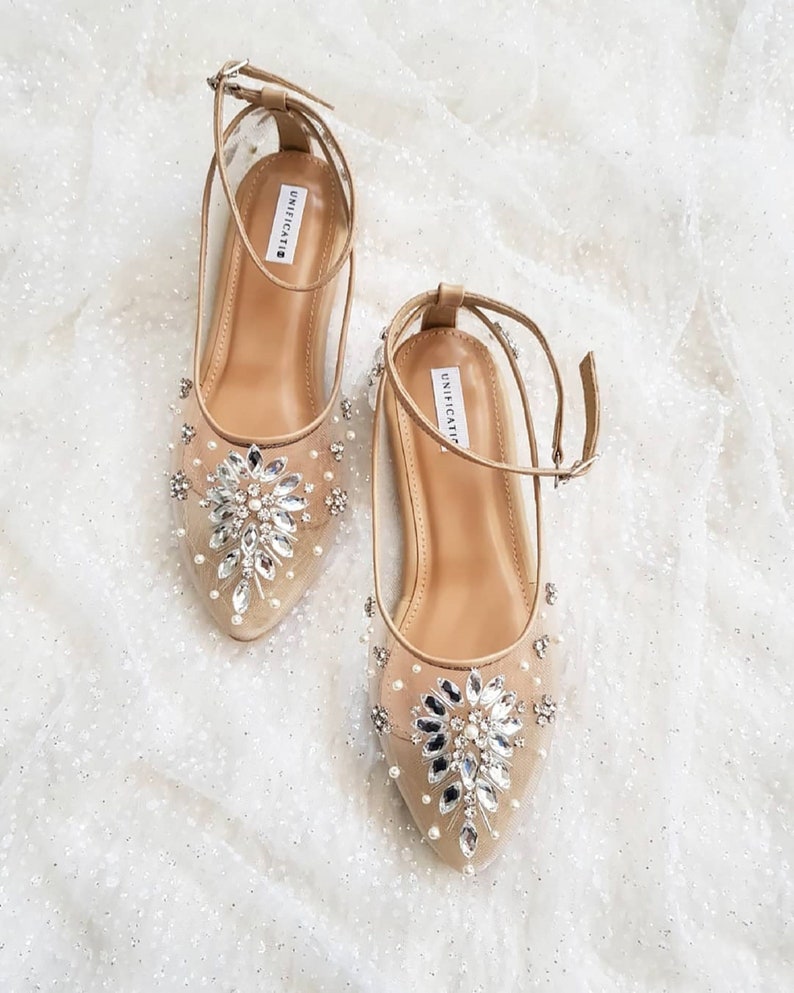 Puede incluir: Un par de zapatos planos de novia beige con correas de tobillo. Los zapatos est&aacute;n adornados con un gran grupo de diamantes de imitaci&oacute;n y perlas en la punta.