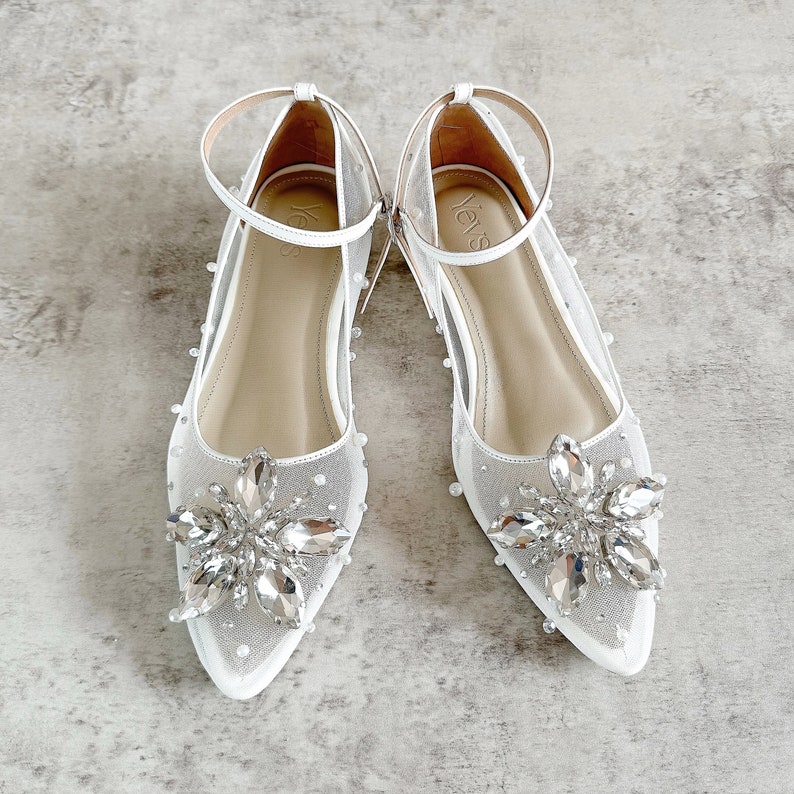 May include: A pair of white pointed-toe flats featuring sheer mesh fabric, ankle straps, and large crystal floral embellishments on the vamps. The shoes are adorned with small white pearls scattered across the mesh and feature the brand name Yevs.
