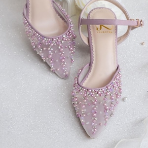 May include: A pair of lavender high-heeled shoes featuring a delicate mesh fabric adorned with pearl-like beads and rhinestones. The shoes have a pointed toe and an ankle strap with a buckle fastening. The brand name "JOOXUTRU" is printed on the insole.