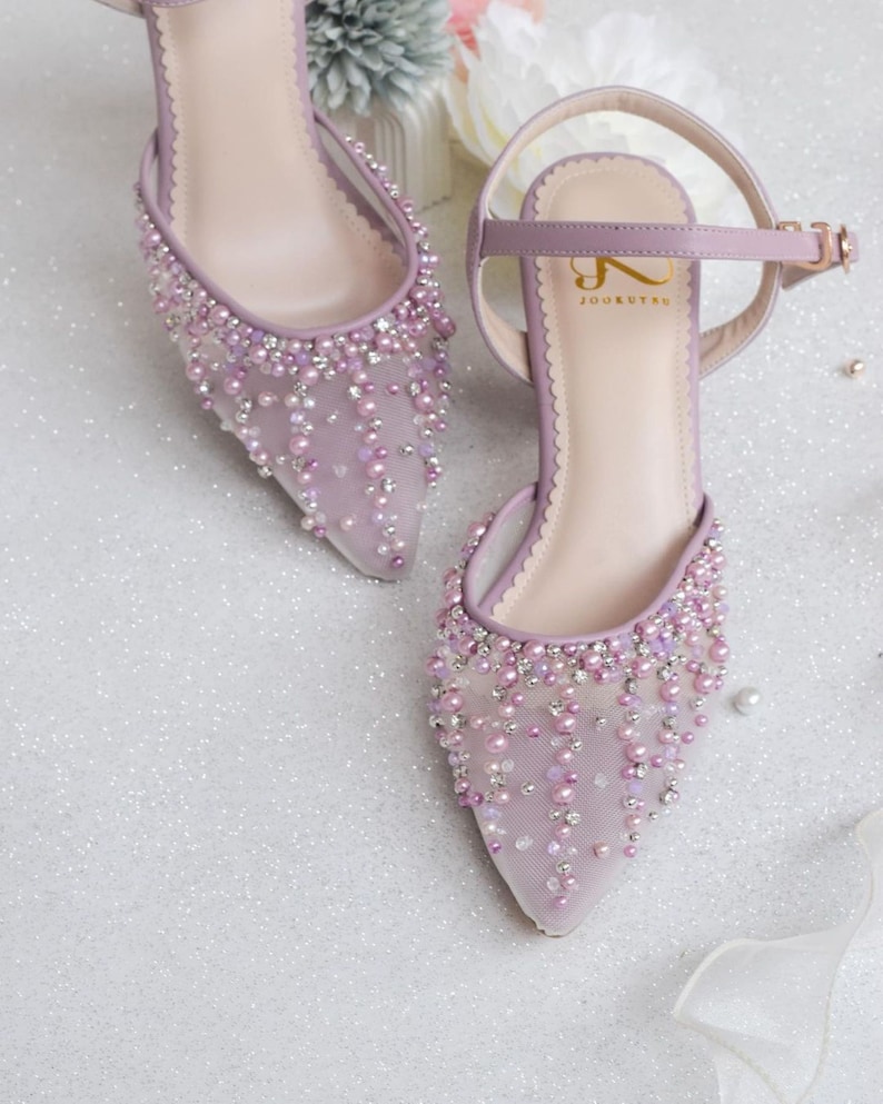 May include: A pair of lavender high-heeled shoes featuring a delicate mesh fabric adorned with pearl-like beads and rhinestones. The shoes have a pointed toe and an ankle strap with a buckle fastening. The brand name "JOOXUTRU" is printed on the insole.