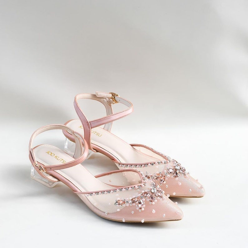 May include: A pair of blush pink bridal heels with a clear block heel and a delicate mesh upper adorned with sparkling rhinestones and pearls. The heels feature an ankle strap with a gold buckle.