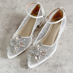 May include: A pair of white pointed-toe flats featuring sheer mesh fabric adorned with small white pearls. Each shoe has a large, multi-faceted clear crystal floral embellishment at the toe and a thin white ankle strap with a silver-toned buckle.