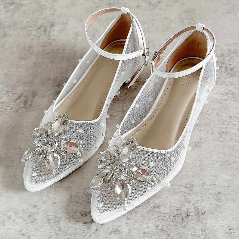 May include: A pair of white pointed-toe flats featuring sheer mesh fabric adorned with small white pearls. Each shoe has a large, multi-faceted clear crystal floral embellishment at the toe and a thin white ankle strap with a silver-toned buckle.