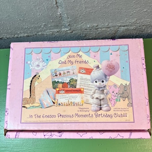 May include: A pink box featuring a Precious Moments figurine holding a heart-shaped balloon. The box has illustrations of cartoon animals, text that reads "Join Me And My Friends...", and the phrase "Happiness is Belonging".