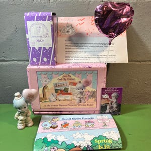 May include: A collection of Precious Moments figurines and related items. The scene includes a purple box, a pink box with the text "Join Me And My Friends", a brochure with the text "Good News Parade", and a heart-shaped balloon.