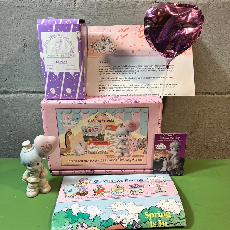 May include: A collection of Precious Moments figurines and related items. The scene includes a purple box, a pink box with the text "Join Me And My Friends", a brochure with the text "Good News Parade", and a heart-shaped balloon.