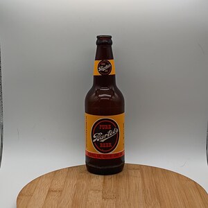 May include: A brown glass beer bottle with a yellow label featuring the text "PURE Bartels BEER" in a circular design. The bottle is on a wooden surface, with a white background.