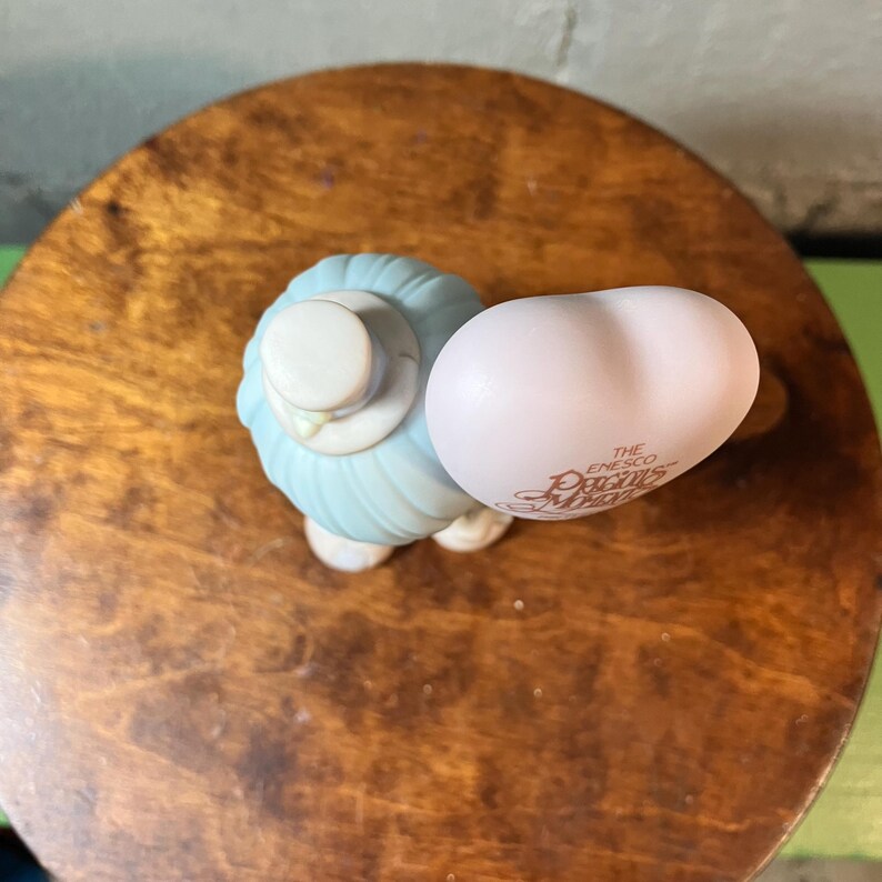 May include: A figurine of a lamb wearing a light blue outfit and a white hat, holding a light pink egg. The egg has the text "The Enesco Precious Moments" printed on it. The figurine is on a wooden surface.