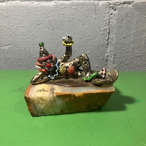 May include: A decorative clown figurine, lying on a stone base. The clown has a red nose, a colorful hat, and a spotted outfit. The base is a light brown color with a polished surface. The figurine is signed "Rome 78".