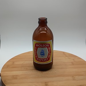May include: A brown glass Molson Export beer bottle with a yellow and white label. The label features the Molson logo, a blue and white design, and the words "EXPORT" and "BIERE". The bottle is on a wooden surface.