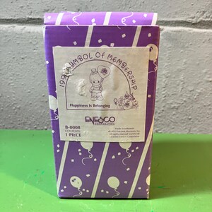 May include: A rectangular purple box with a white and purple balloon pattern. The box features a label with the text "1993 Symbol of Membership" and "Happiness Is Belonging". The Enesco Corporation logo is also visible.