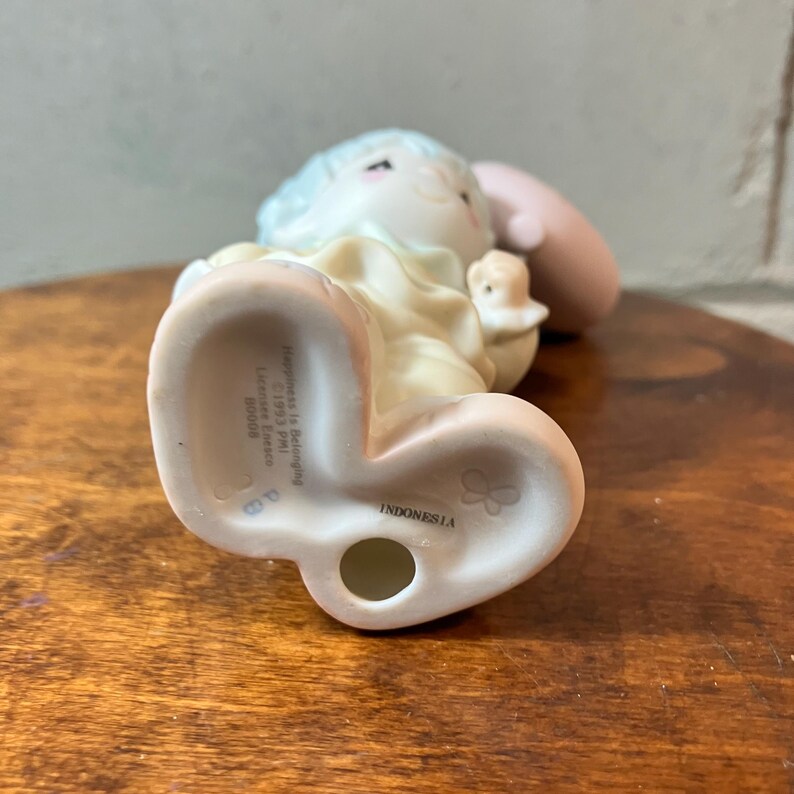 May include: A ceramic figurine of a child holding a small bird. The figurine has a light blue hair, a yellow dress, and a pink cheek. The base of the figurine has the text "Happiness Is Belonging" and "Indonesia".