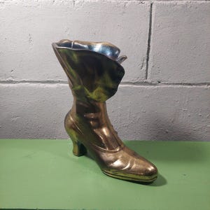 May include: A vintage-style brass boot-shaped vase. The boot has a high heel and a flared top, with a metallic gold and green finish. The vase is likely used for decorative purposes.