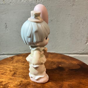 May include: A porcelain figurine of a child holding a pink balloon. The child wears a light blue hat and has light blue hair. The outfit is cream-colored with pink polka dots. The figurine is on a wooden surface.