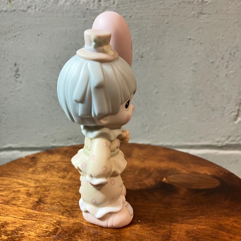 May include: A porcelain figurine of a child holding a pink balloon. The child wears a light blue hat and has light blue hair. The outfit is cream-colored with pink polka dots. The figurine is on a wooden surface.