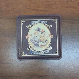 May include: A vintage Hershey's Milk Chocolate tin. The rectangular tin is brown with a decorative border. The center features an oval illustration of a man and woman. The words "Hershey's", "Pure", "Milk", and "Chocolate" are visible.