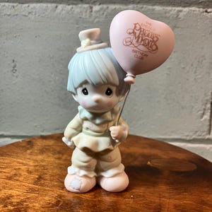 May include: A Precious Moments figurine of a clown holding a pink heart-shaped balloon. The figurine has light blue hair and a matching hat. The balloon has the words "The Precious Moments Birthday Club" printed on it.