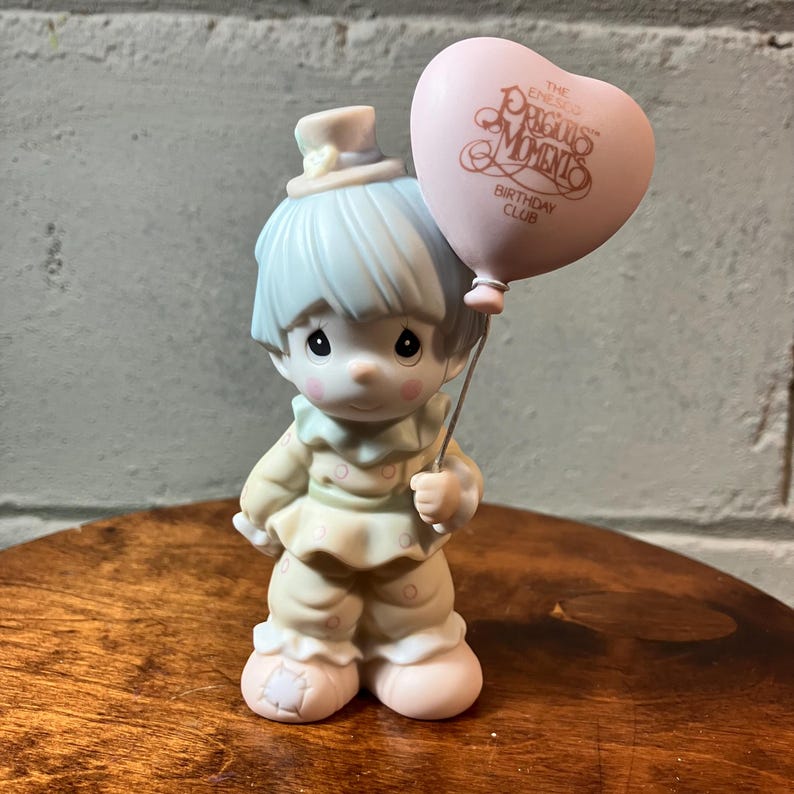 May include: A Precious Moments figurine of a clown holding a pink heart-shaped balloon. The figurine has light blue hair and a matching hat. The balloon has the words "The Precious Moments Birthday Club" printed on it.