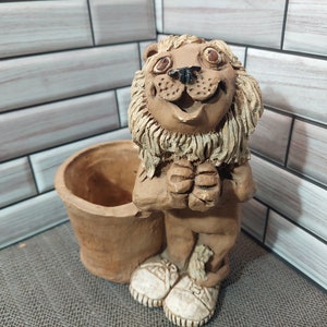 May include: A brown ceramic lion figurine with a wide smile and a planter pot. The lion is holding its paws together in front of its chest. The planter pot is brown and has a wide opening.