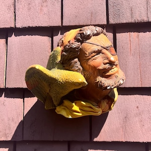 May include: A detailed, colorful ceramic pirate head with an eye patch and a parrot. The pirate has a brown beard and hair, and the parrot is yellow and green. The sculpture is mounted on a brown surface.