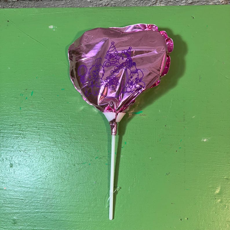 May include: A pink heart-shaped balloon on a white stick. The balloon has a purple illustration of a clown and other characters. The balloon is made of a shiny, metallic material, and the stick is white.