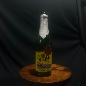 May include: A green glass beer bottle with a yellow label featuring the text "DINKEL ACKER" and an illustration of a horse-drawn carriage. The bottle is sealed with a silver foil and sits on a wooden stool.