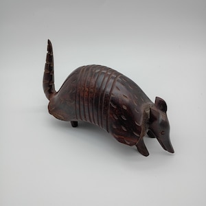 May include: A dark brown, hand-carved wooden armadillo figurine. The sculpture features a segmented shell design, a pointed snout, small ears, and a raised, textured tail. The wood grain is visible throughout the polished, smooth surface.