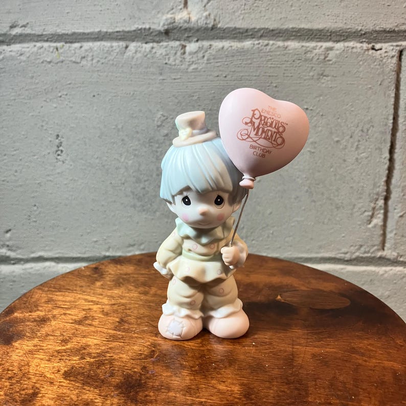 May include: A Precious Moments figurine of a young boy holding a pink heart-shaped balloon. The boy has light blue hair and is wearing a cream-colored outfit. The balloon has the words "Precious Moments Birthday Club" printed on it.