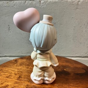 May include: A figurine of a child holding a pink heart-shaped balloon. The child wears a light blue hat and has light blue hair. The child's outfit is cream-colored with pink polka dots. The figurine is on a wooden surface.