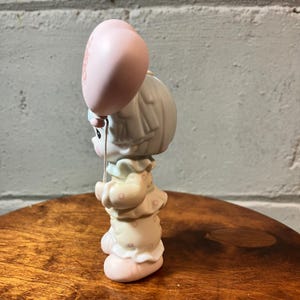 May include: A ceramic figurine of a child holding a pink balloon. The child is wearing a light yellow dress with pink polka dots and a ruffled collar. The figurine has a sweet, innocent expression and is a collectible item.