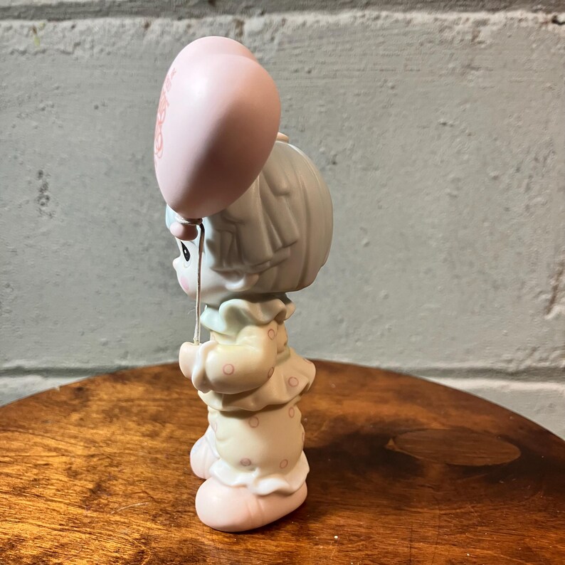 May include: A ceramic figurine of a child holding a pink balloon. The child is wearing a light yellow dress with pink polka dots and a ruffled collar. The figurine has a sweet, innocent expression and is a collectible item.
