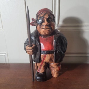 May include: A wooden figurine of a pirate with a red bandana, black eye patch, and a black jacket. The pirate is holding a wooden stick in his right hand.