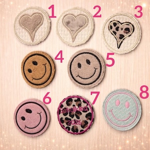 Könnte beinhalten: Eine Sammlung von acht gestickten Aufnähern. Die Designs umfassen Herzen, Smiley-Gesichter und ein Leopardenmuster. Die runden Aufnäher sind in verschiedenen Farben erhältlich, darunter Braun, Rosa und Blau. Einige Aufnäher tragen den Text "Schulkind 2026".