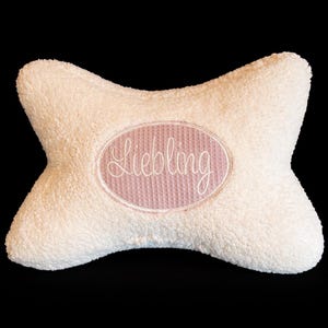 Könnte beinhalten: Ein knochenförmiges, cremefarbenes Kissen mit einer weichen, strukturierten Oberfläche. Ein ovales Feld in der Mitte zeigt das gestickte Wort "Liebling" in zarter Schrift auf rosa Hintergrund. Das Kissen ist für Komfort konzipiert.
