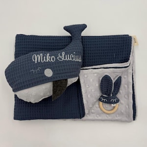 May include: A navy blue baby activity book with a whale and bunny design. The whale has the name "Miko Lucius" embroidered on it. The bunny has a wooden ring and is attached to a gray, textured fabric.