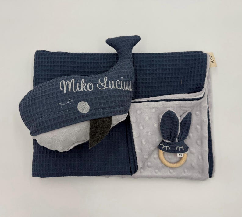 May include: A navy blue baby activity book with a whale and bunny design. The whale has the name "Miko Lucius" embroidered on it. The bunny has a wooden ring and is attached to a gray, textured fabric.