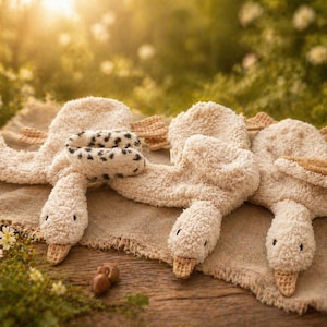 Könnte beinhalten: Drei Plüsch-Schwan-Spielzeuge in Creme- und Beigetönen sind auf einer strukturierten beigefarbenen Oberfläche angeordnet. Ein Schwan hat einen schwarz-weiß gefleckten Hals. Die Schwäne haben strukturierte Körper, beige Flügel und Waffel-Textur Schnäbel. Der Hintergrund ist eine verschwommene Außenszene.