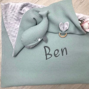 May include: A baby blanket and accessories in shades of gray and green. The blanket has a waffle texture and the name "Ben" embroidered on it. A whale-shaped plush toy and a wooden teether with a bunny face are included.
