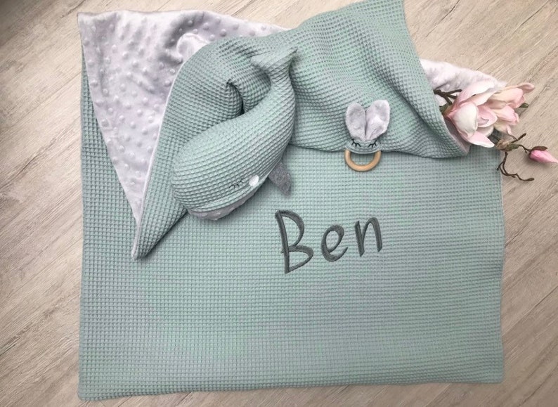 May include: A baby blanket and accessories in shades of gray and green. The blanket has a waffle texture and the name "Ben" embroidered on it. A whale-shaped plush toy and a wooden teether with a bunny face are included.