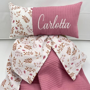 Könnte beinhalten: Eine rosa-weiße Babydecke mit Waffelstruktur und Blumenmuster. Die Decke ist in der Mitte gefaltet und hat ein Kissen mit dem Namen "Carlotta" bestickt.